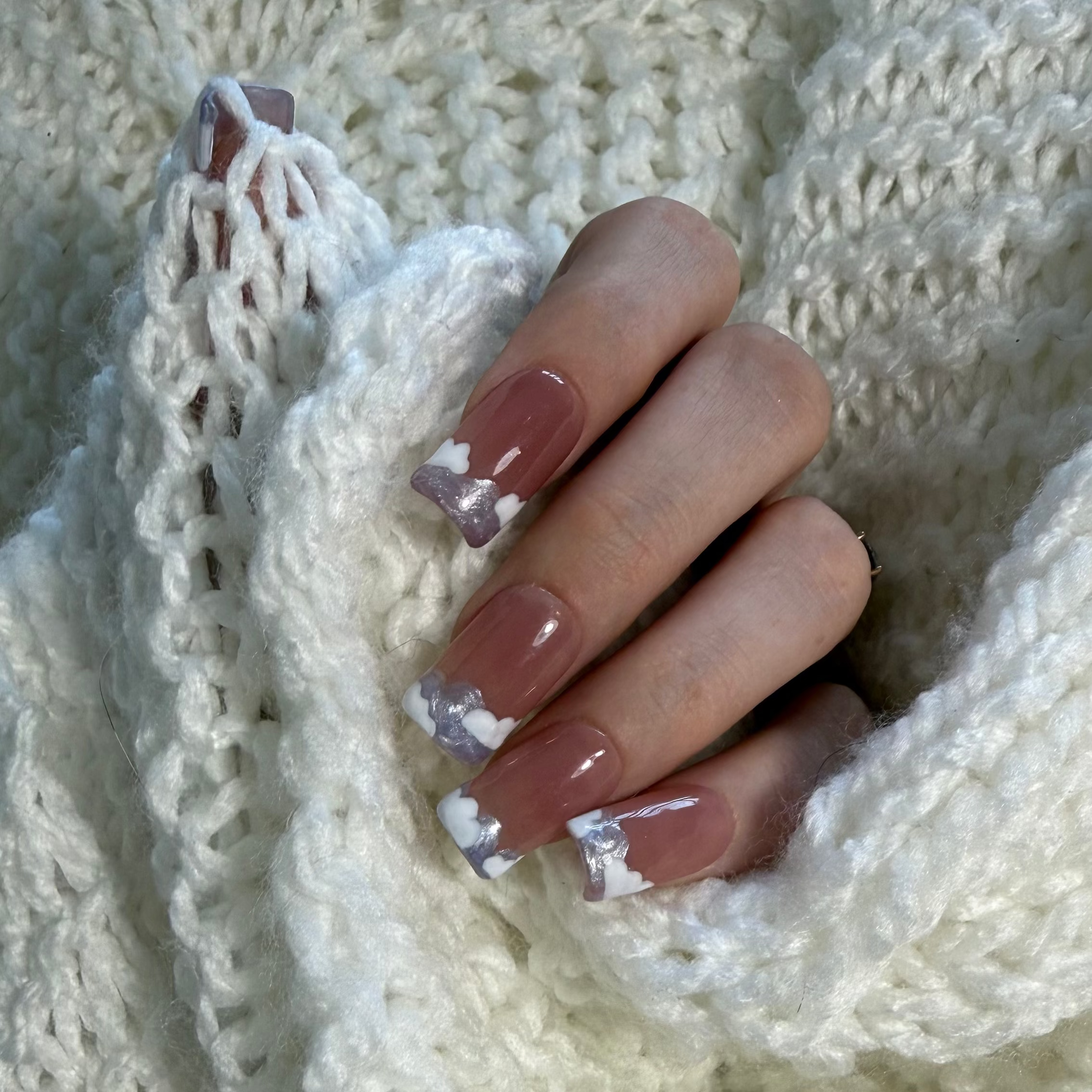 Pink base nails with a purple swirly French tip and white clouds on top on a hand wrapped in a crocheted sweater