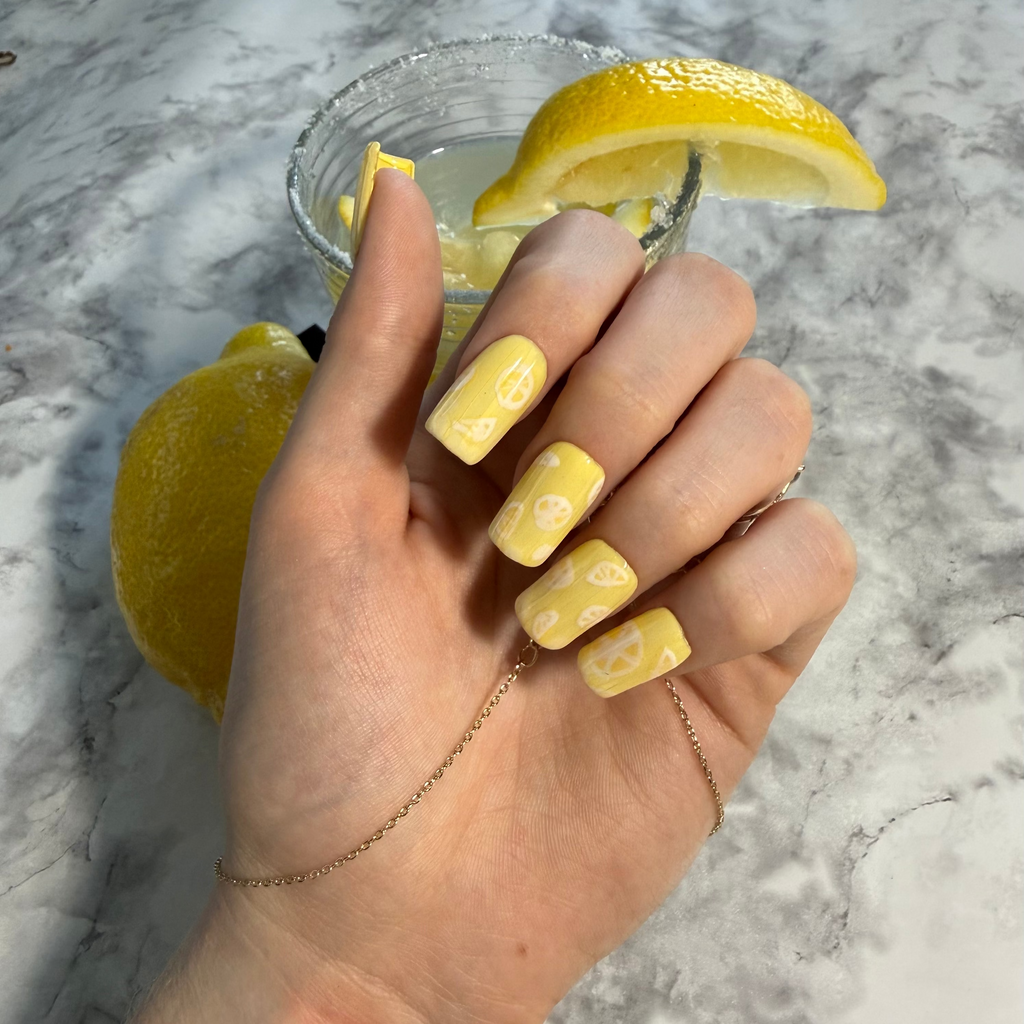 Yellow nail art designs with lemon slices with a ribbed glass effect on top on a hand with a glass of lemonade in the background
