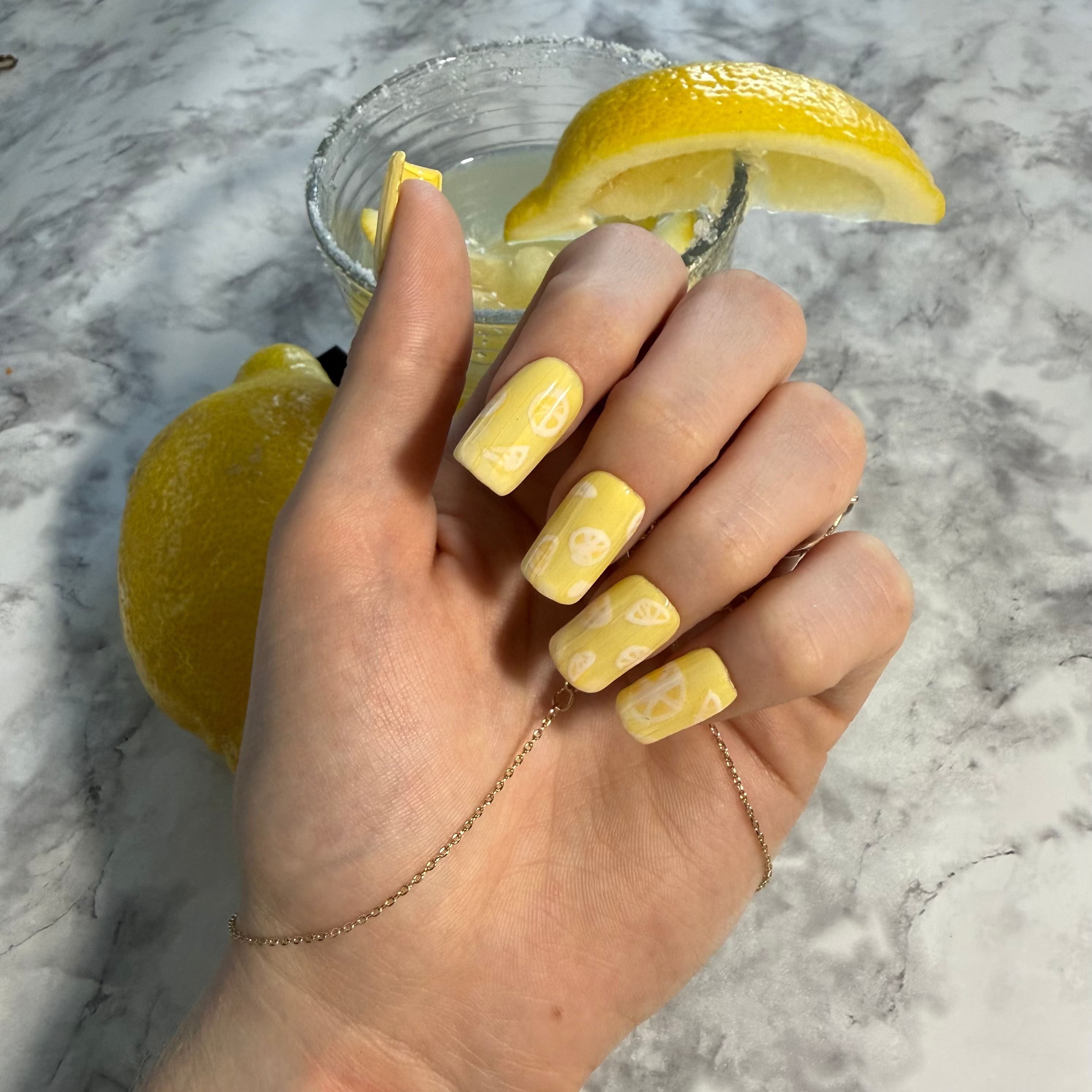 Yellow nail art designs with lemon slices with a ribbed glass effect on top on a hand with a glass of lemonade in the background