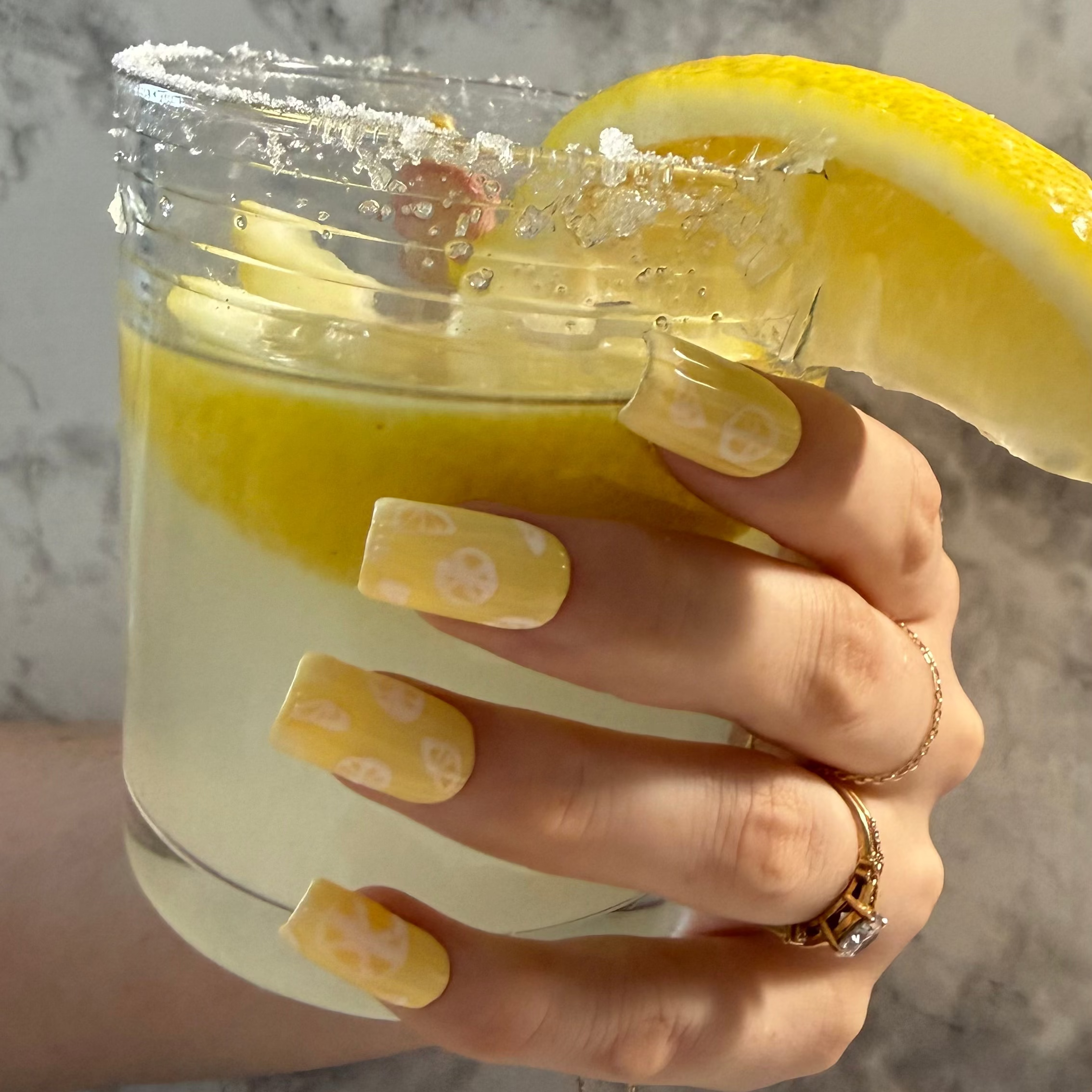 Yellow nail art designs with lemon slices with a ribbed glass effect on top on a hand holding a glass of lemonade with a lemon slice and sugar on top