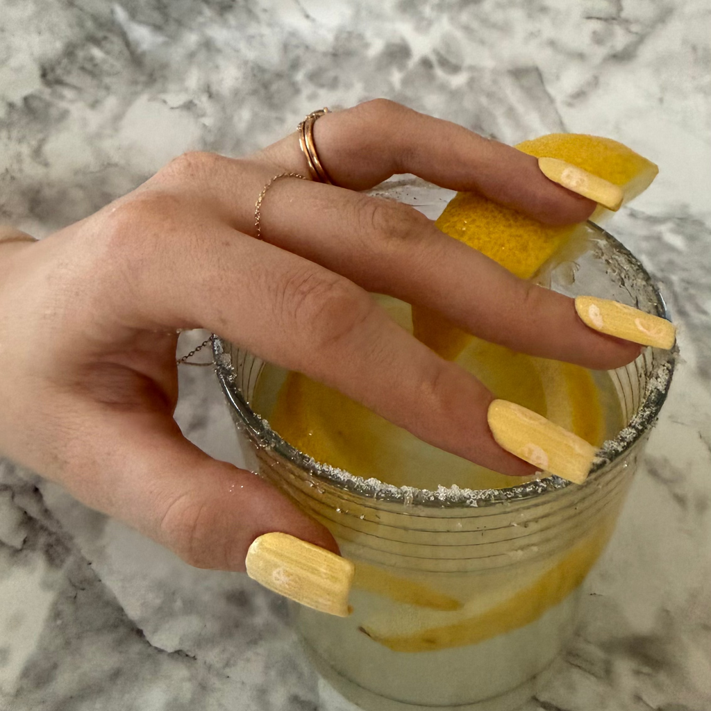 Yellow nail art designs with lemon slices with a ribbed glass effect on top on a hand that is placed on a glass of lemonade with a lemon slice and sugar on top