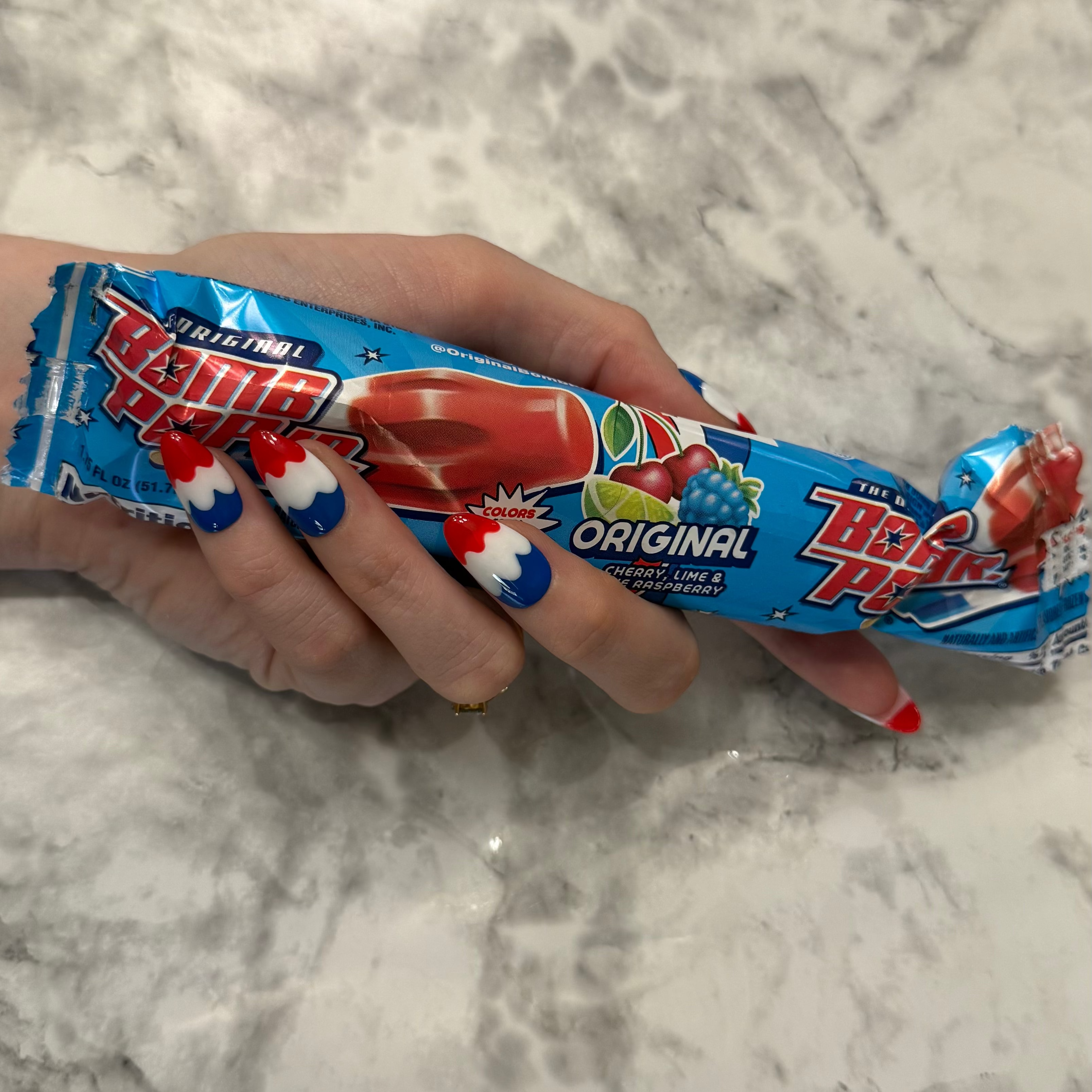 Nail art with red, white, and blue design that is a swirly to imitate a melting bomb pop on a hand holding a bomb pop wrapper