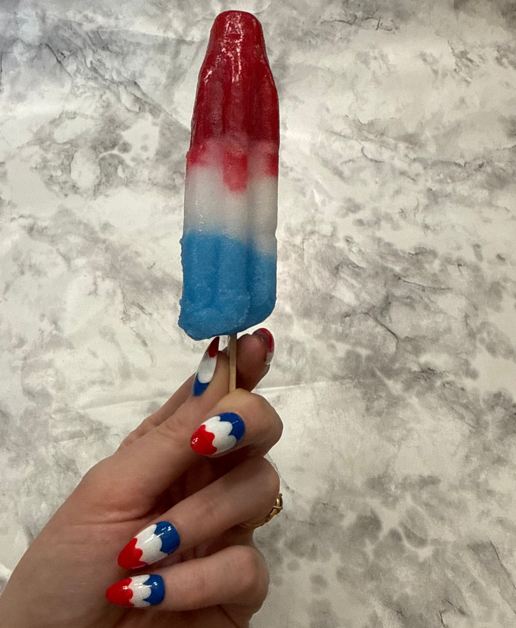 Nail art with red, white, and blue design that is a swirly to imitate a melting bomb pop on a hand holding a melting bomb pop