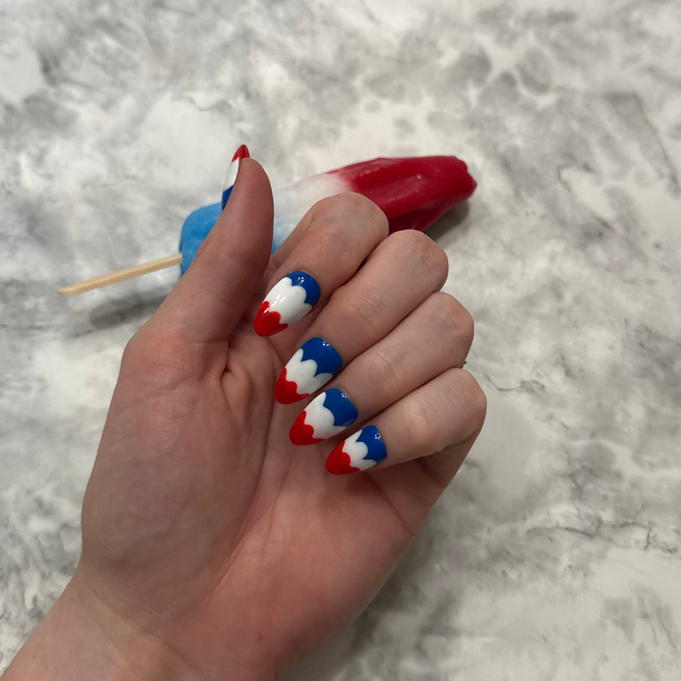 Nail art with red, white, and blue design that is a swirly to imitate a melting bomb pop on a hand in front of a bomb pop