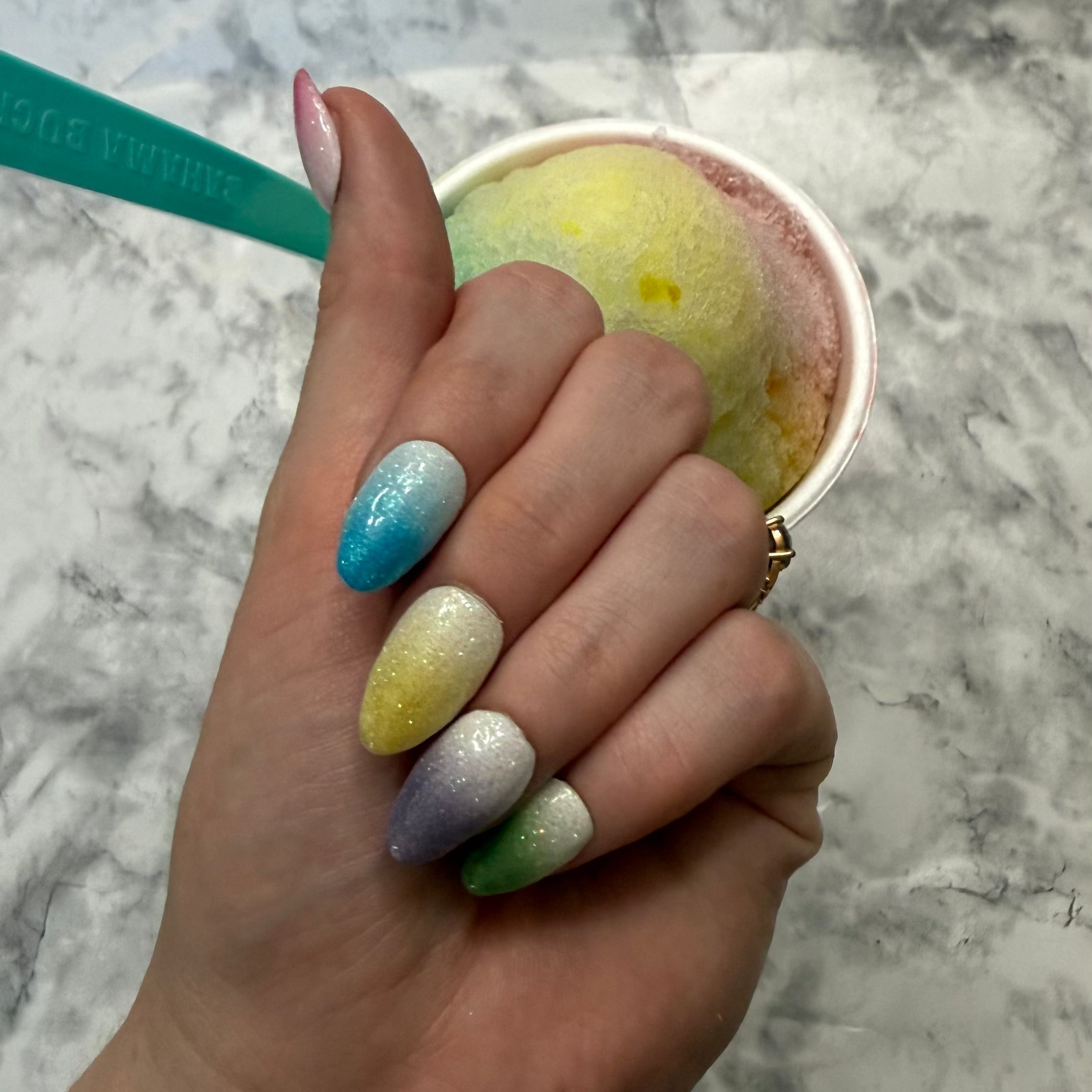 Five glittery almond shaped nails that are pink, blue, yellow, purple, and green made to look like snow cones on a hand in front of a snow cone that is rainbow colored with a blue spoon in it