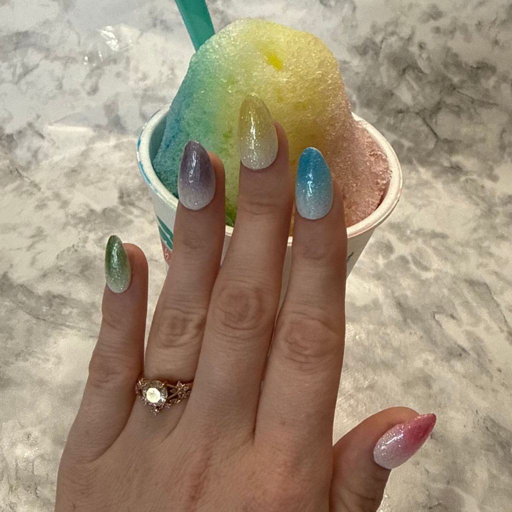 Five glittery almond shaped nails that are pink, blue, yellow, purple, and green made to look like snow cones on a hand in front of a rainbow snow cone with a blue spoon
