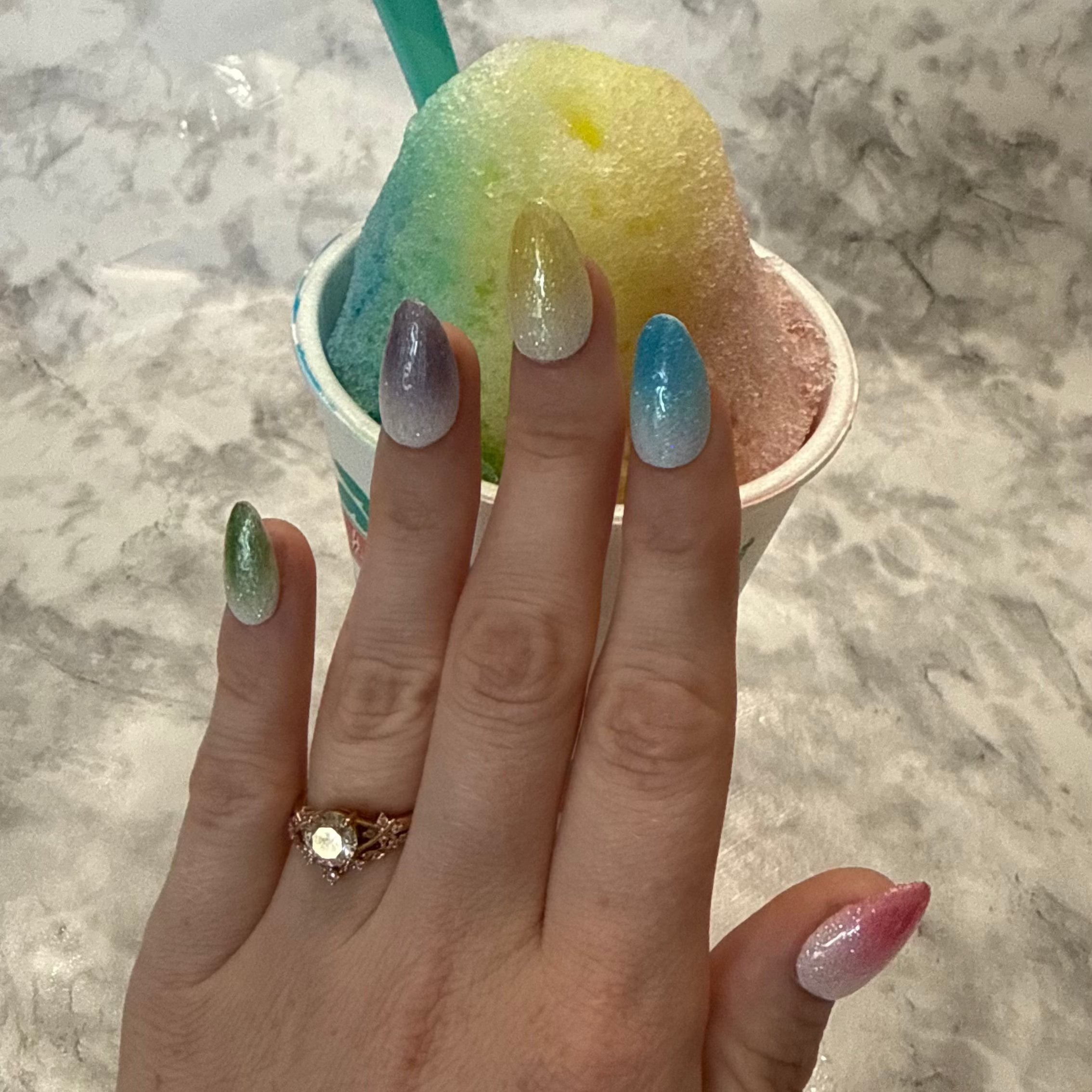 Five glittery almond shaped nails that are pink, blue, yellow, purple, and green made to look like snow cones on a hand in front of a rainbow snow cone with a blue spoon