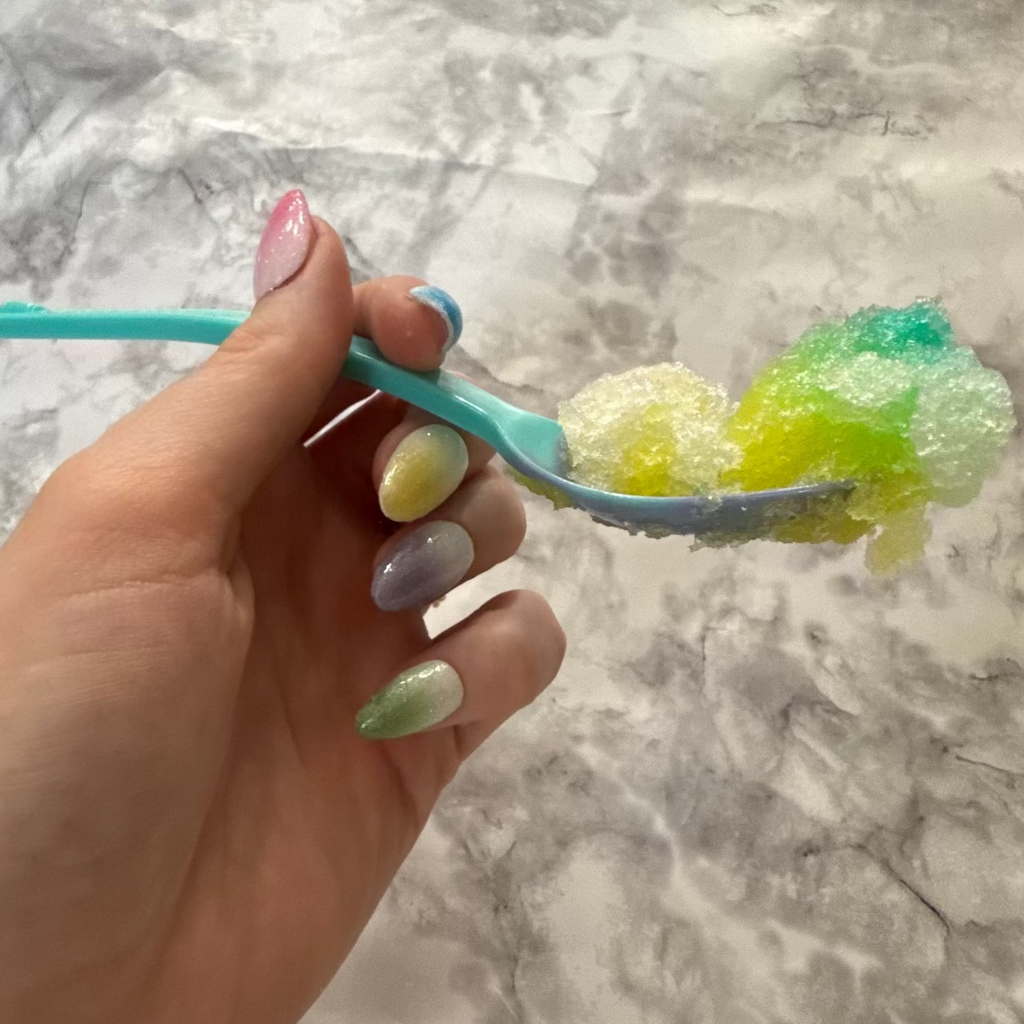 Five glittery almond shaped nails that are pink, blue, yellow, purple, and green made to look like snow cones on a hand holding a spoonful of snow cone that is yellow and green and blue on a blue spoon
